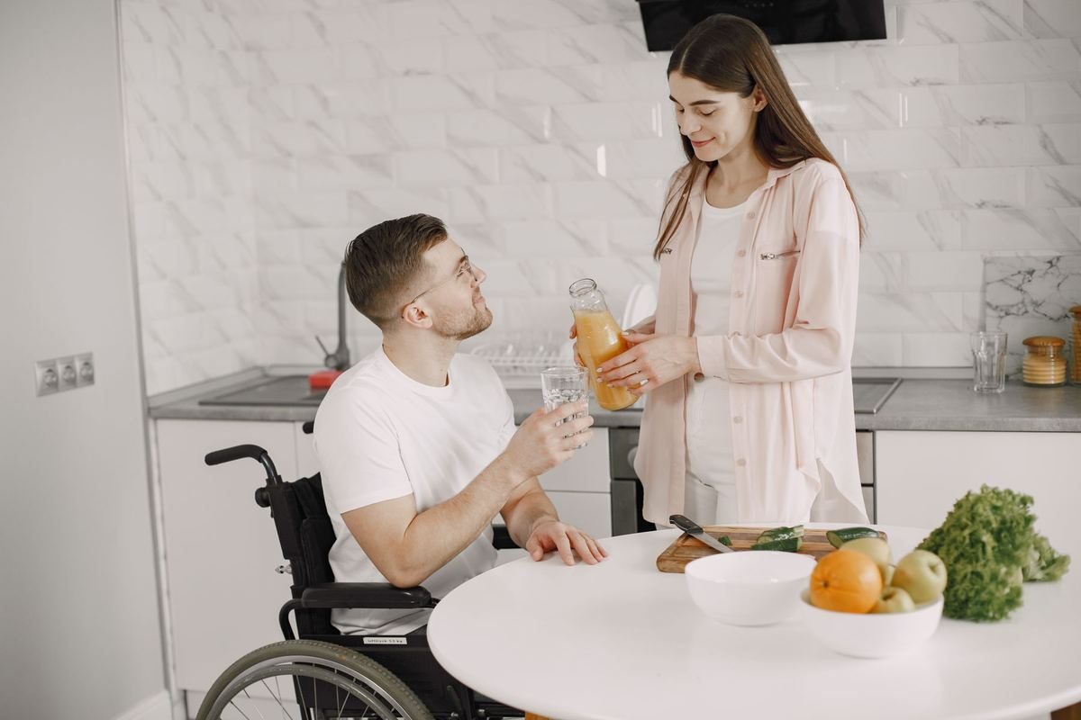 A young man who uses a wheelchair at his kitchen table at home, with a carer bringing him a drink