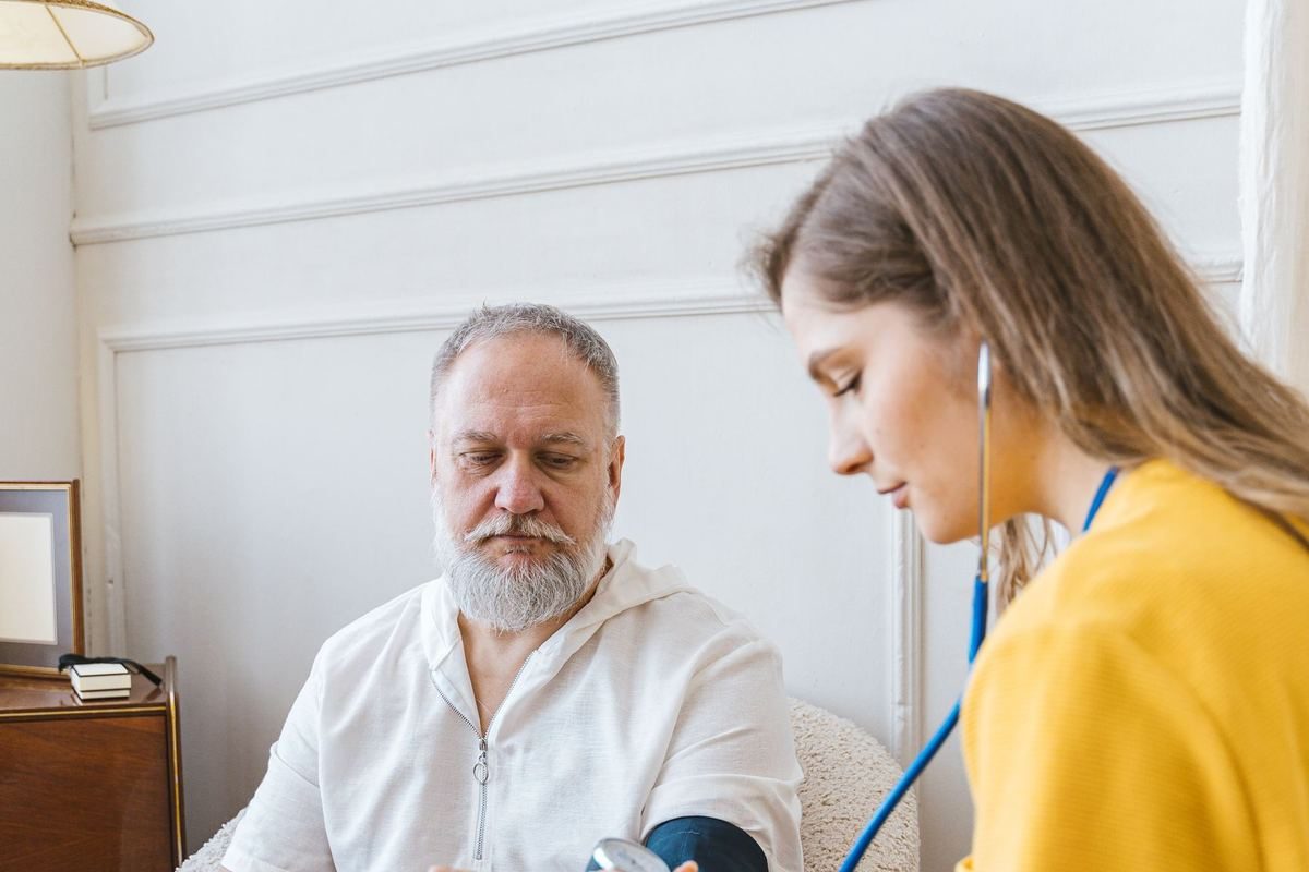 A carer in a tunic checking an older man's blood pressure in his living room