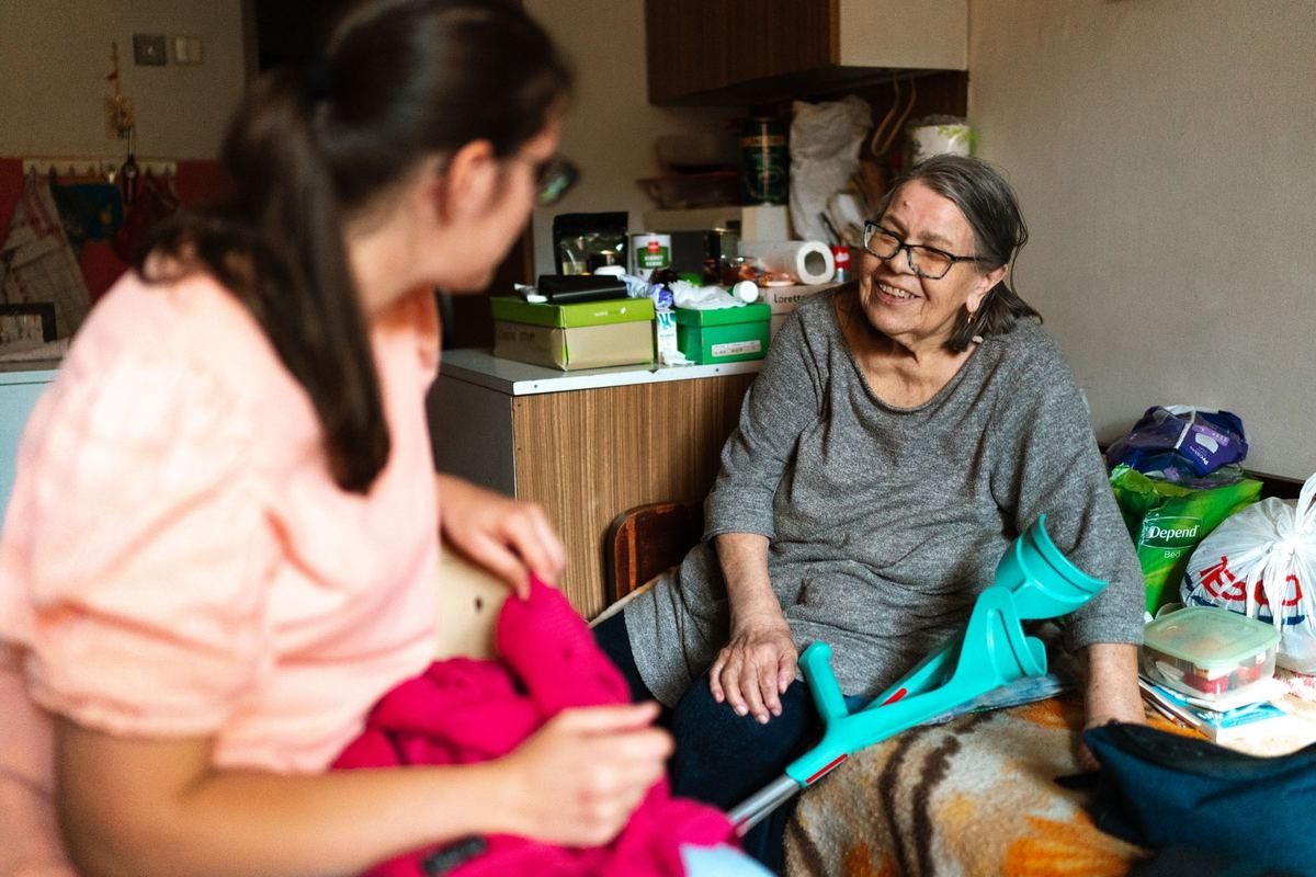 A carer sitting with a smiling older woman using crutches at home, helping her unpack