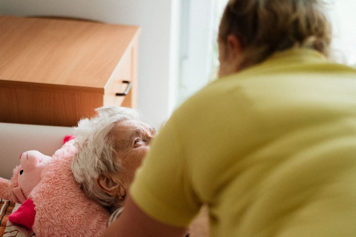 A carer leaning over to check on an older woman resting in bed